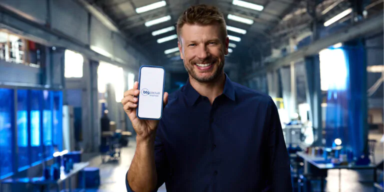 Rodrigo Hilbert sorridente segura um celular exibindo a marca "BTG Pactual Empresas", dentro de um galpão industrial iluminado. A imagem promove os serviços do banco voltados para empresas.